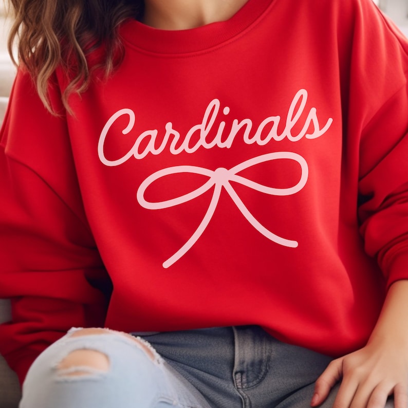 May include: Red sweatshirt with the word "Cardinals" in white script and a pink bow underneath.