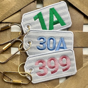 May include: Three embroidered keychains with white tags and gold-colored hardware. The tags display the numbers "1A" in green, "30A" in blue, and "399" in pink. The tags have a striped pattern.