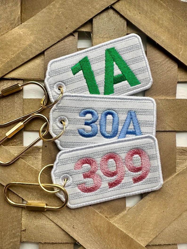 May include: Three embroidered keychains with white tags and gold-colored hardware. The tags display the numbers "1A" in green, "30A" in blue, and "399" in pink. The tags have a striped pattern.