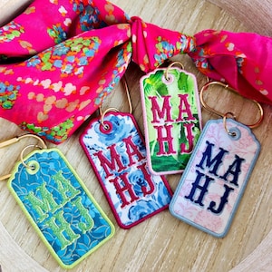 May include: Four embroidered keychains with the initials "MA HJ" in various colors, including blue, green, and pink, are displayed. A bright pink floral scarf is knotted in the background. The keychains are on a wooden tray.