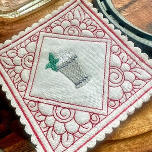 May include: Embroidered white coaster with a red floral design and scalloped edges. The center features an embroidered image of a beverage with whipped cream and a mint leaf. The coaster is square and rests on a wooden surface.