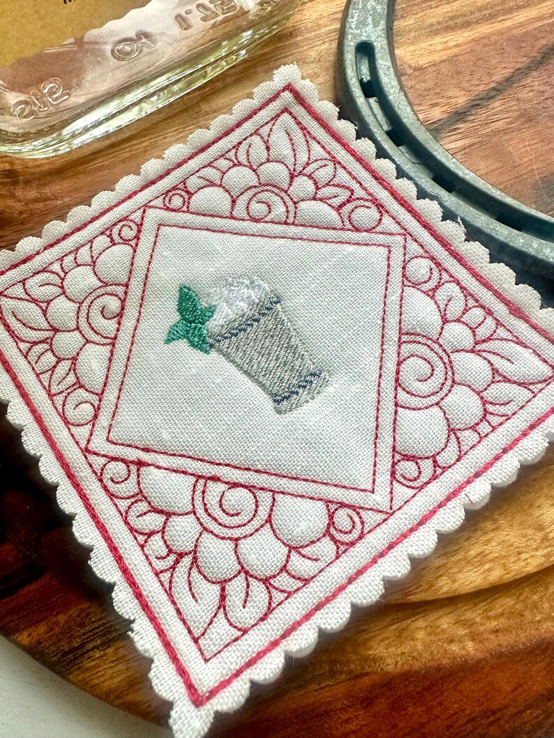May include: Embroidered white coaster with a red floral design and scalloped edges. The center features an embroidered image of a beverage with whipped cream and a mint leaf. The coaster is square and rests on a wooden surface.