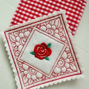 May include: Embroidered white square coaster with a red rose in the center, surrounded by a red decorative border. The coaster has a scalloped edge and is placed on a red and white checkered fabric.