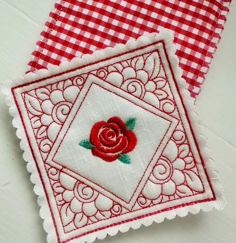 May include: Embroidered white square coaster with a red rose in the center, surrounded by a red decorative border. The coaster has a scalloped edge and is placed on a red and white checkered fabric.