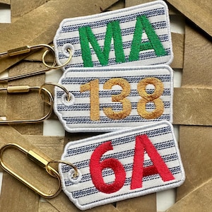 May include: Three embroidered keychains with white and blue striped backgrounds. The top keychain has the letters "MA" in green. The middle keychain has the numbers "138" in gold. The bottom keychain has the letters "6A" in red. Each keychain has a gold-colored clasp.