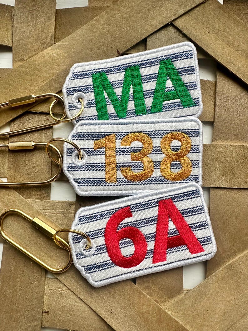 May include: Three embroidered keychains with white and blue striped backgrounds. The top keychain has the letters "MA" in green. The middle keychain has the numbers "138" in gold. The bottom keychain has the letters "6A" in red. Each keychain has a gold-colored clasp.