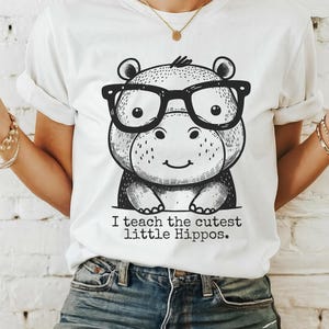 May include: White t-shirt with a black and white illustration of a hippo wearing glasses. The text "I teach the cutest little Hippos." is printed below the illustration.