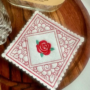 May include: A square, white embroidered coaster with a red rose in the center. The coaster has a scalloped edge and a red floral design around the rose. The coaster is on a wooden surface.