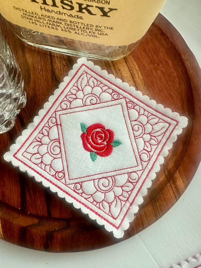 May include: A square, white embroidered coaster with a red rose in the center. The coaster has a scalloped edge and a red floral design around the rose. The coaster is on a wooden surface.