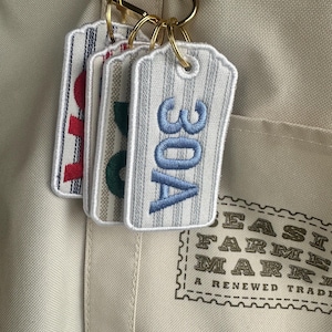 May include: Embroidered fabric tags with vertical stripes and gold key rings. The tags display the numbers "30A" in blue, "80" in green, and "10" in red. The tags are attached to a beige tote bag.