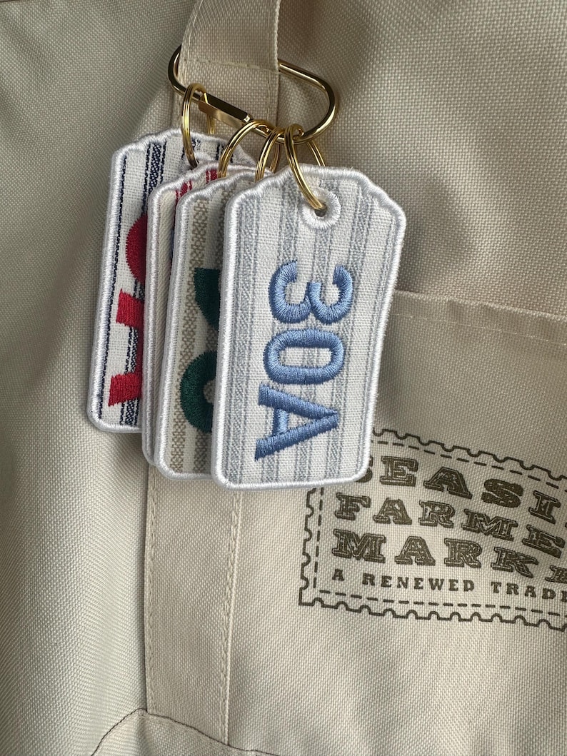 May include: Embroidered fabric tags with vertical stripes and gold key rings. The tags display the numbers "30A" in blue, "80" in green, and "10" in red. The tags are attached to a beige tote bag.