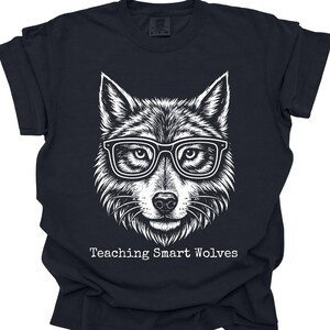 Puede incluir: Camiseta negra con una ilustración blanca detallada de un lobo con gafas. El texto "Teaching Smart Wolves" está impreso debajo. La camiseta está hecha de un material suave.