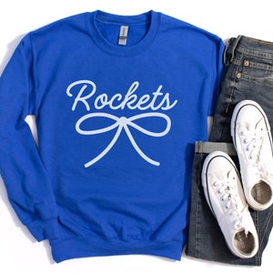 May include: Royal blue sweatshirt with white text that reads "Rockets" and a white bow. The sweatshirt is laying on a white surface with a pair of blue jeans and white sneakers.