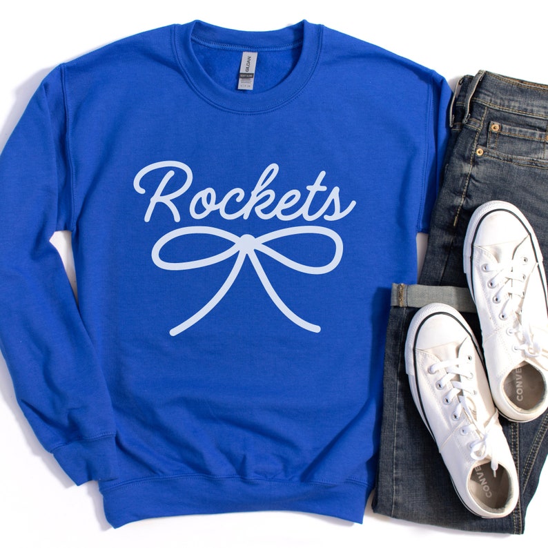 May include: Royal blue sweatshirt with white text that reads "Rockets" and a white bow. The sweatshirt is laying on a white surface with a pair of blue jeans and white sneakers.