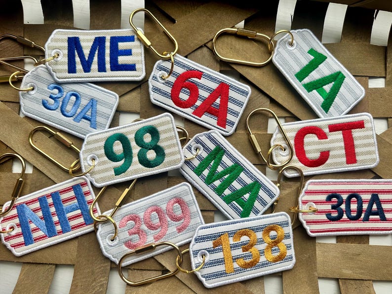 May include: A collection of embroidered keychains with various state abbreviations and numbers. The keychains feature a striped background with different colored text and gold-tone hardware.