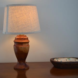 May include: A wooden table lamp with a beige lampshade and a brown, carved wooden base. The lamp is lit, casting a warm glow. A wooden bowl with decorative elements sits nearby.