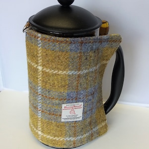 Könnte beinhalten: Eine schwarze und gläserne French Press Kaffeemaschine mit einer gelben und blauen karierten Tweed-Stoffhülle. Die Hülle trägt ein Etikett mit der Aufschrift "Harris Tweed" mit einem Logo und dem Text "Outer Hebrides of Scotland 100% Pure New Wool".