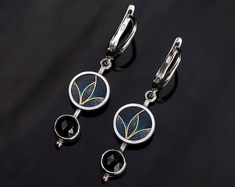 May include: A pair of silver earrings with black and green enamel accents. Each earring features a round pendant with a stylized leaf design and a smaller black gemstone pendant.