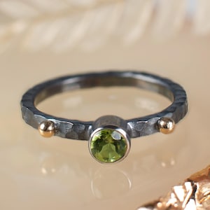 May include: A silver ring with a textured band and a round, light green gemstone. The ring has two small gold accents on either side of the gemstone. The ring is displayed on a light-colored surface.