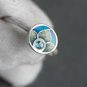 May include: A silver ring with a round, floral-inspired design. The ring features a central, light blue gemstone surrounded by a blue and gray enamel pattern. The ring is held against a gray background.