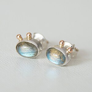 May include: A pair of silver stud earrings with a blue-green labradorite gemstone and three small gold accents on each earring.