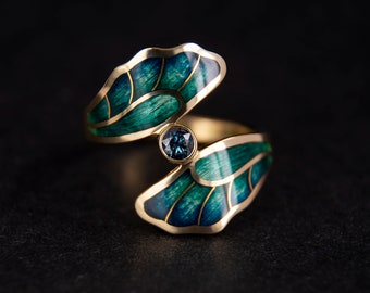 Puede incluir: Un anillo abierto de oro con dos secciones en forma de hoja rellenas de esmalte azul verdoso y verde con vetas doradas. Una piedra preciosa redonda de color azul oscuro está engastada en el centro entre los dos motivos de hojas.