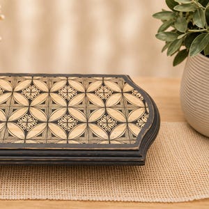 May include: A rectangular wooden tray with a black distressed frame and a light wood surface featuring a repeating geometric pattern of black etched circles and floral motifs. The tray has rounded corners and a smooth, flat top surface.