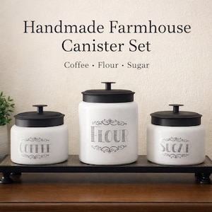 May include: A handmade farmhouse canister set with three white ceramic jars with black lids. The jars are labeled "Coffee", "Flour", and "Sugar" in a decorative font. The set is displayed on a black tray.