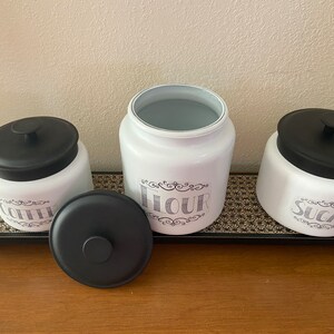 May include: A set of three white ceramic kitchen canisters with black lids, labeled "Coffee", "Flour", and "Sugar" in a decorative font. The canisters sit on a black tray with a gold floral pattern, ideal for kitchen storage.