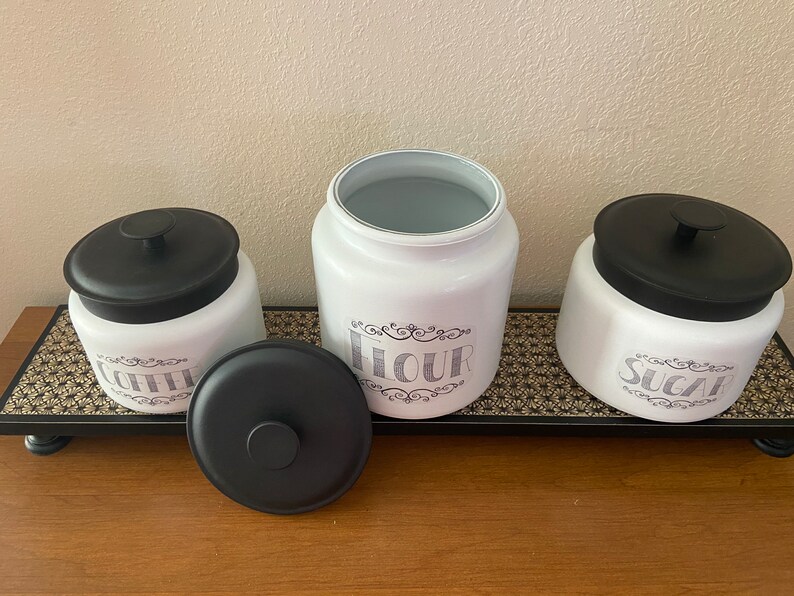 May include: A set of three white ceramic kitchen canisters with black lids, labeled "Coffee", "Flour", and "Sugar" in a decorative font. The canisters sit on a black tray with a gold floral pattern, ideal for kitchen storage.