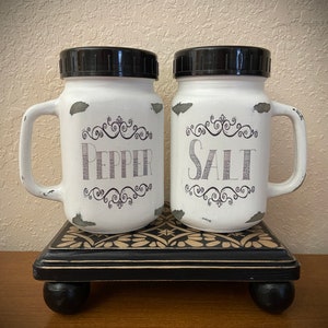 May include: White ceramic salt and pepper shakers with black lids and handles. The shakers are decorated with black script that says "Salt" and "Pepper" with decorative flourishes. The shakers are sitting on a black and white patterned wooden stand with black feet.