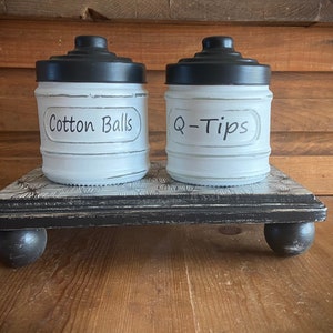 May include: Two white painted jars with black lids on a black wooden stand with black feet. The jars are labeled "Cotton Balls" and "Q-Tips".