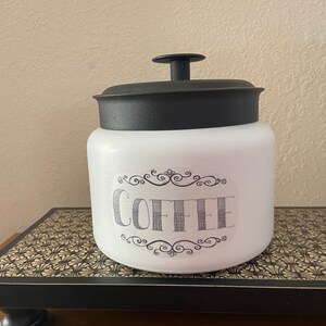 May include: White ceramic coffee canister with a black lid and handle. The word "COFFEE" is printed on the front in a decorative font, framed by ornate scrollwork. The canister is sitting on a decorative black and gold tray.