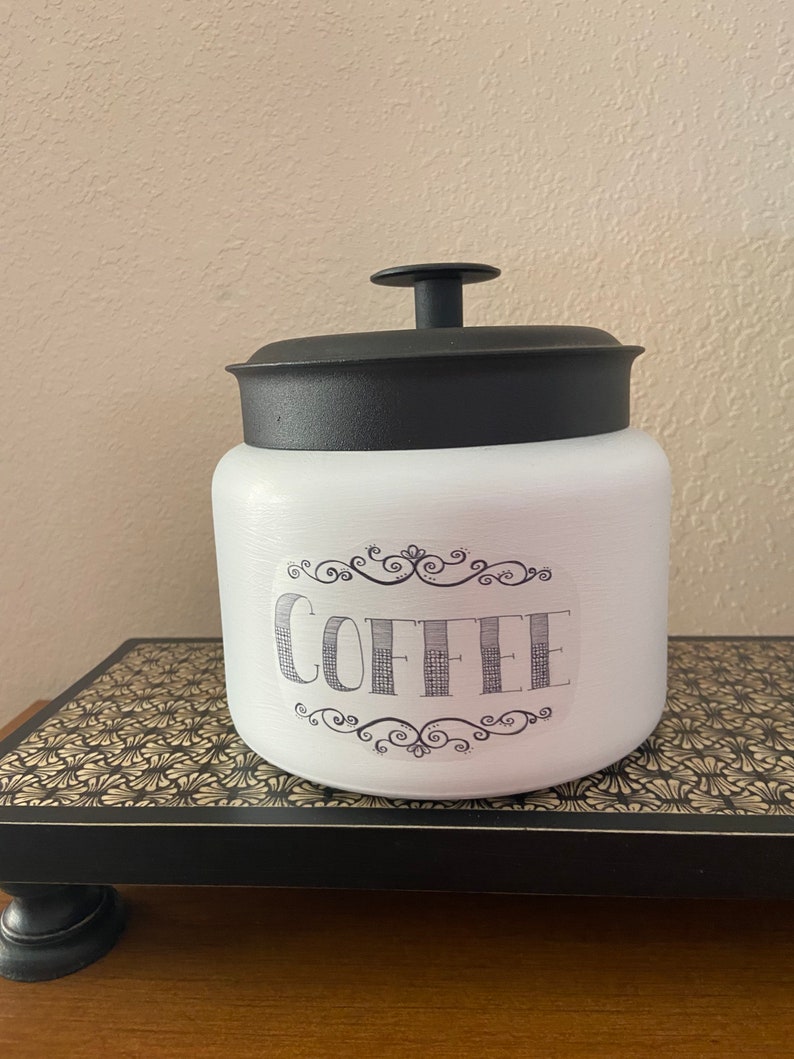 May include: White ceramic coffee canister with a black lid and handle. The word "COFFEE" is printed on the front in a decorative font, framed by ornate scrollwork. The canister is sitting on a decorative black and gold tray.