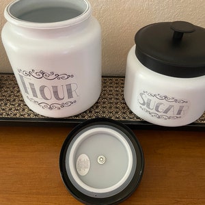 May include: White kitchen canisters labeled "Flour" and "Sugar" with black lids. The flour canister is open, revealing a gray interior. The sugar canister has a black lid. The text is in a decorative font.