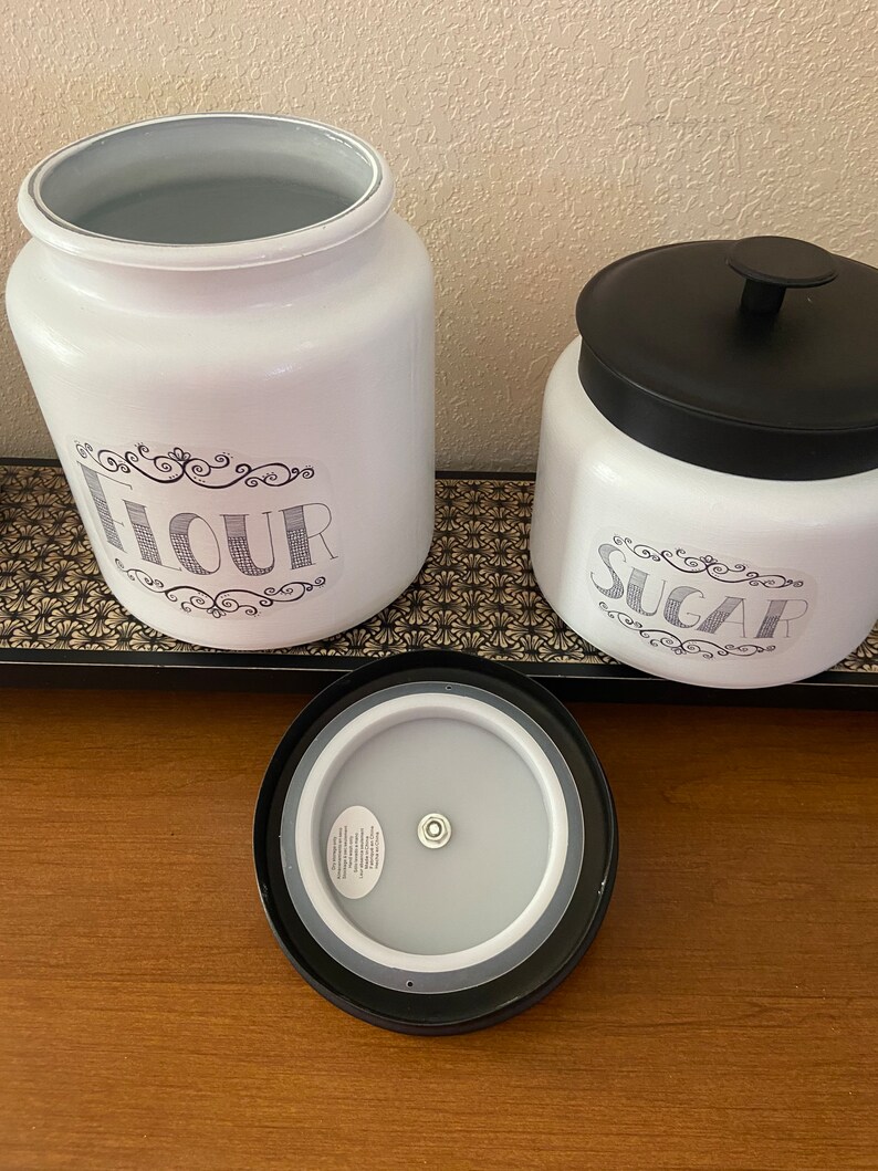 May include: White kitchen canisters labeled "Flour" and "Sugar" with black lids. The flour canister is open, revealing a gray interior. The sugar canister has a black lid. The text is in a decorative font.