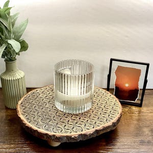 May include: A decorative wooden tray with a geometric pattern holds a clear glass candle holder with a lit candle. A small framed picture of a sunset and a green vase with greenery are also on the wooden surface.
