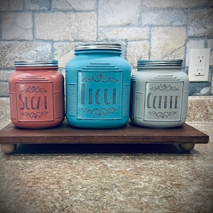 May include: Three square glass canisters with silver lids, each featuring a decorative black label with scrollwork. The left canister is coral pink and labeled SUGAR, the center is bright blue and labeled FLOUR, and the right is light gray and labeled COFFEE.