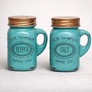 May include: A pair of turquoise ceramic salt and pepper shakers in a mason jar design. Each shaker has a copper-colored lid and a handle. The text "Pepper" and "Salt" are displayed on the front.