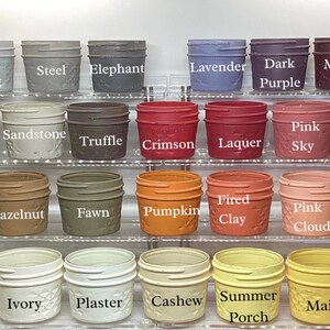 May include: A collection of small, decorative jars in various colors, including silver, lavender, and crimson. Each jar has a textured pattern and a label with the color name. The jars are arranged on clear shelves, showcasing a wide range of hues.