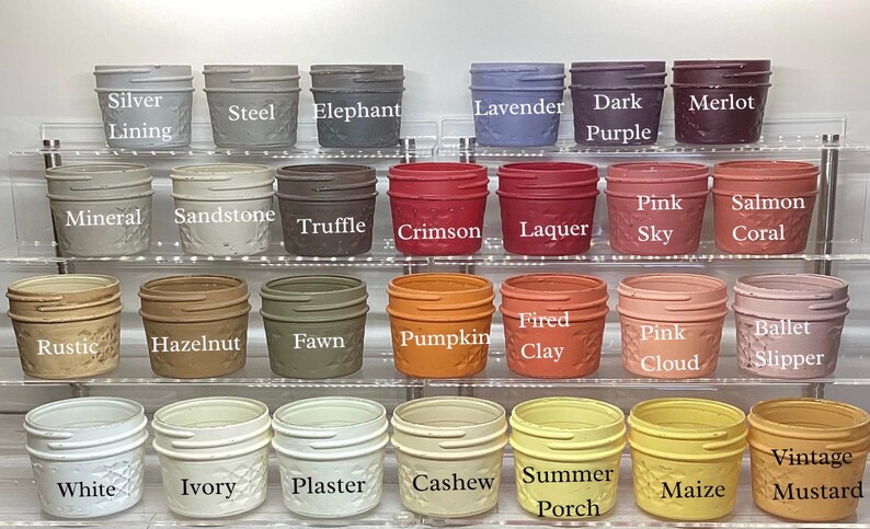 May include: A collection of small, decorative jars in various colors, including silver, lavender, and crimson. Each jar has a textured pattern and a label with the color name. The jars are arranged on clear shelves, showcasing a wide range of hues.