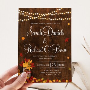 May include: Rustic wedding invitation with a woodgrain background, featuring the names "Sarah Daniels" and "Richard O'Brien" in white script. The invitation includes fall foliage, a mason jar with leaves, and string lights.