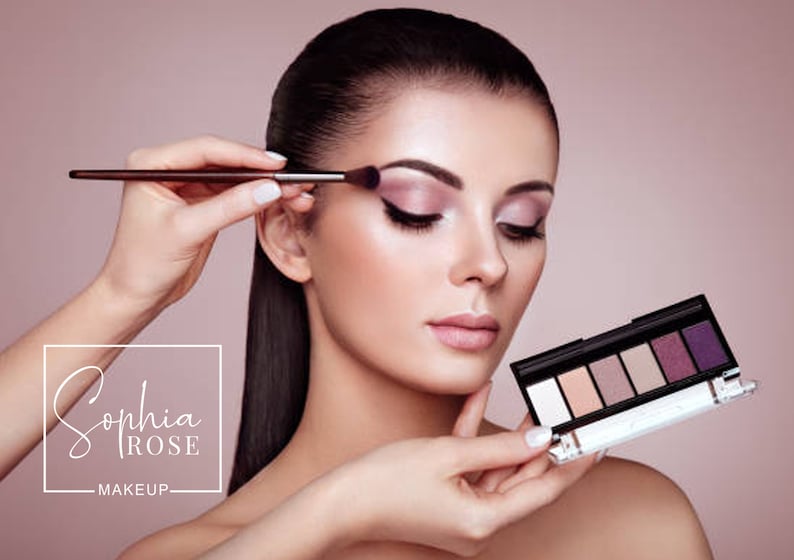 May include: A woman is applying eyeshadow with a brush. She is holding a makeup palette with a variety of shades. The palette has a black case with a silver trim. The brand name "Sophia Rose Makeup" is visible in the lower left corner of the image.