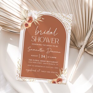 May include: A rectangular invitation card with the text "bridal shower" in a decorative arch shape. The card features floral and leaf accents, with the bride's name and event details. The colour scheme includes terracotta, white, and gold.