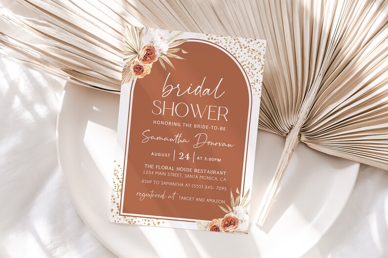 May include: A rectangular invitation card with the text "bridal shower" in a decorative arch shape. The card features floral and leaf accents, with the bride's name and event details. The colour scheme includes terracotta, white, and gold.