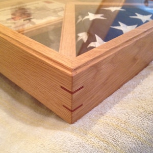 May include: Wooden display case with a glass top, showcasing a folded American flag and other memorabilia. The case is made of light-colored wood with a mitered corner joint reinforced with red accents. The flag is blue with white stars.