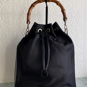 May include: Black bucket bag with a bamboo handle and a drawstring closure. The bag has a silver chain strap and silver hardware.