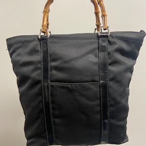 May include: Black tote bag with a bamboo handle. The bag has a rectangular shape with a black fabric exterior and a bamboo handle. The handle is attached to the bag with silver-colored metal hardware and black leather straps.
