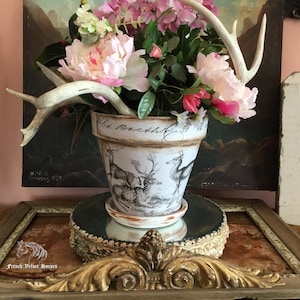 May include: A white ceramic planter with a deer design and the text "Wild Orchids" is filled with pink and white flowers and topped with antlers. The planter is sitting on a gold decorative stand.