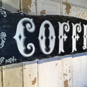 May include: A black wooden sign with white lettering that reads "COFFEE" in a decorative font. The sign has a distressed look with white paint chipping off the black surface. The sign is decorated with white scrollwork.